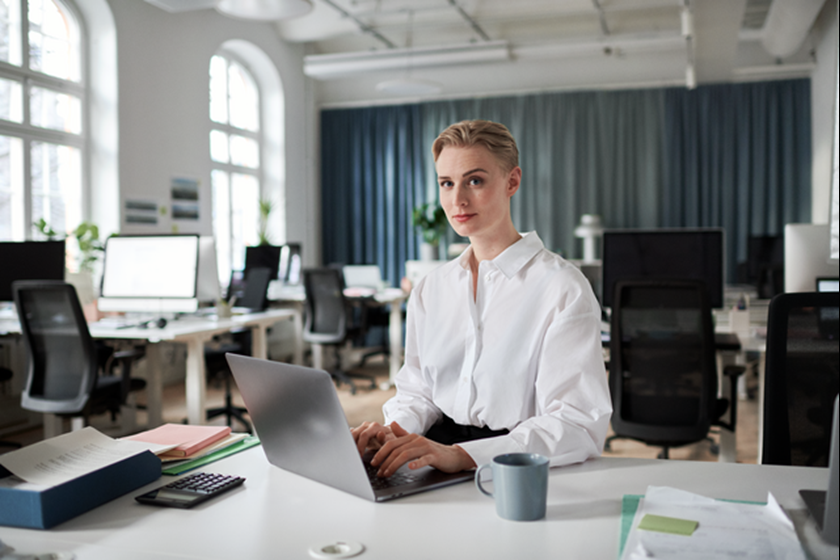 Zelfverzekerde vrouw met witte blouse achter haar laptop op klein kantoor