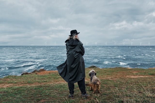 Man in zwarte jas met hoed en hond in de wind aan zee