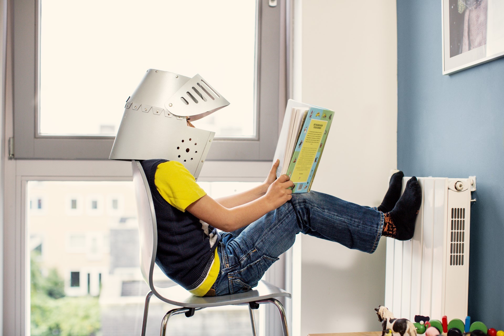 Jongetje met zelfgemaakte helm leest een spannend boek met zijn voeten tegen de radiator 