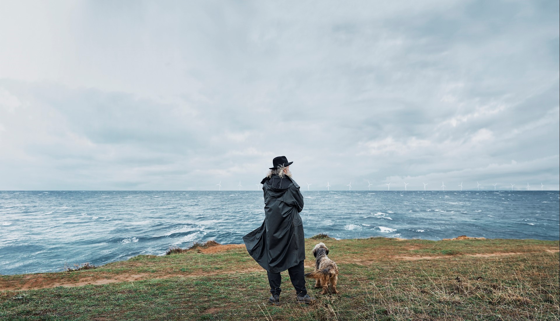Oude man in zwarte jas met hond in de wind aan de kust 