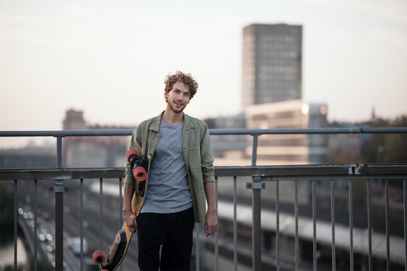 Jongeman staat lachend op een brug met zijn skateboard onder zijn arm
