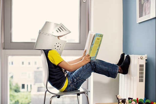Jongetje met zelfgemaakte helm leest een spannend boek met zijn voeten tegen de radiator 