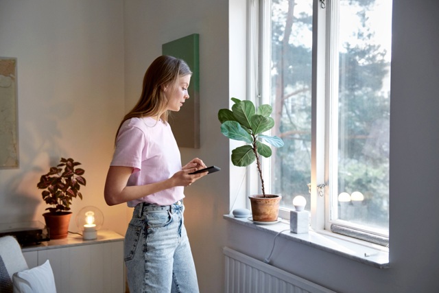 Jonge vrouw in roze shirt kijkt uit het raam met mobiel in de hand